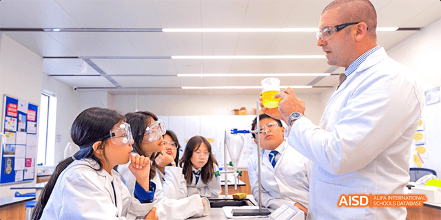 A group of students from Harrow LiDe School Haikou are gathered around a teacher in a lab, watching a science experiment.