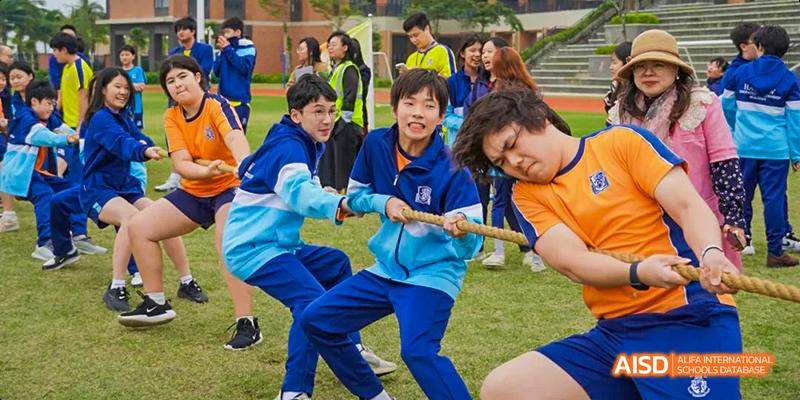 A group of students from Harrow LiDe School Haikou competes in a tug-of-war game on a grassy field.