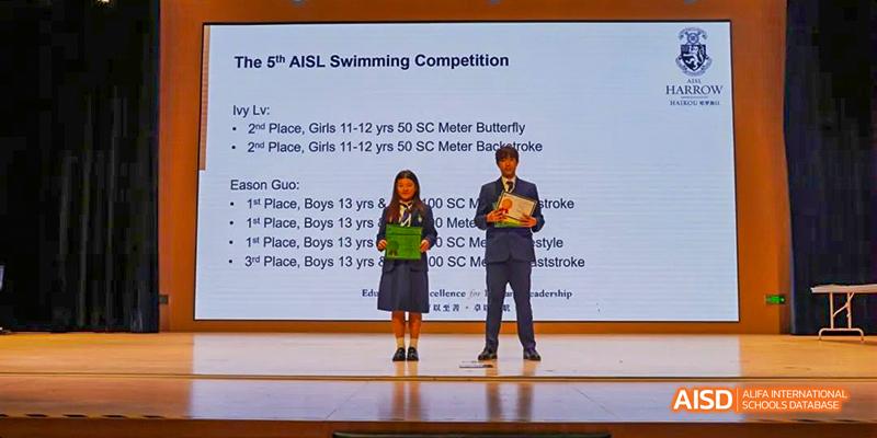 Two students from Harrow LiDe School Haikou stand on a stage receiving awards or certificates for a swimming competition.