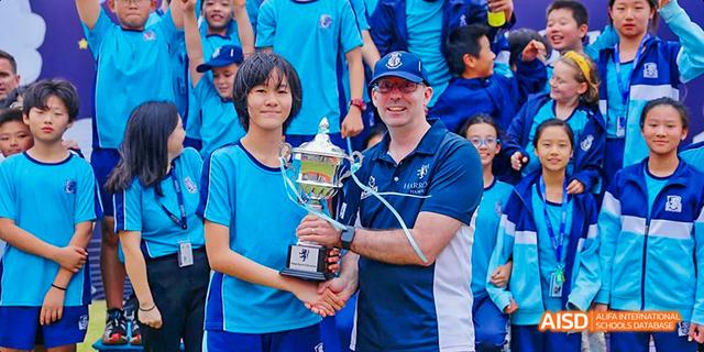 A male student from Harrow LiDe School Haikou and a coach pose together, holding a large trophy.