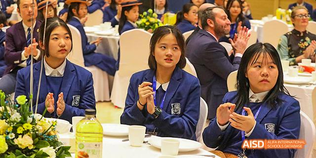 A female student from Harrow LiDe School Haikou in a school uniform is seated at a table and appears to be listening to a presentation.