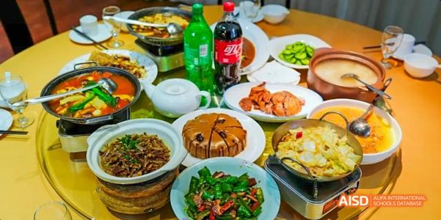 A table is set with a variety of Chinese dishes, including a cake and two large bottles of soda, showing a meal scene at Harrow LiDe School Haikou.