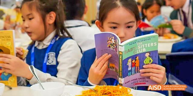 A young female student from Harrow LiDe School Haikou reads a book while eating a bowl of noodles.