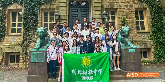 A large group of students and teachers from the International Department of Chengdu Shude High School standing in front of a classical building and holding the school's green banner.