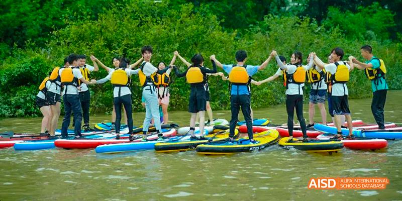Students from the International Department of Chengdu Shude High School engaging in outdoor water sports, holding hands while standing on paddleboards on a lake.