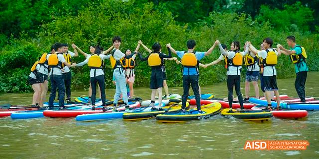 Students from the International Department of Chengdu Shude High School engaging in outdoor water sports, holding hands while standing on paddleboards on a lake.
