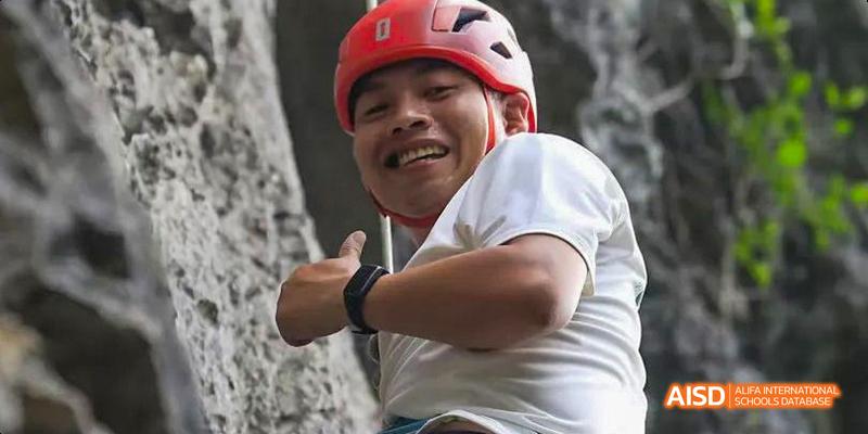 A smiling male student from the International Department of Chengdu Shude High School wearing a red climbing helmet and giving a thumbs up while rock climbing.