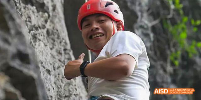 A smiling male student from the International Department of Chengdu Shude High School wearing a red climbing helmet and giving a thumbs up while rock climbing.