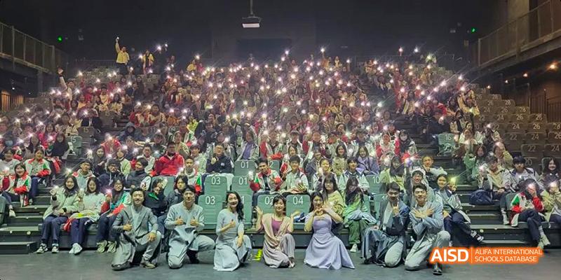 A large audience holding up cell phone lights during an event organized by the International Department of Chengdu Shude High School, with a small group of performers kneeling in the foreground.