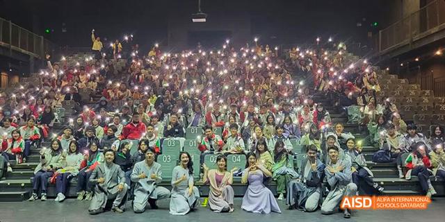 A large audience holding up cell phone lights during an event organized by the International Department of Chengdu Shude High School, with a small group of performers kneeling in the foreground.
