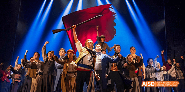 A vibrant stage performance by students and actors from the International Department of Chengdu Shude High School, with one male performer holding a large red banner.