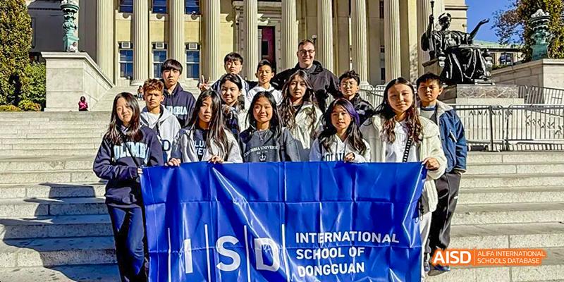 A diverse group of International School of Dongguan students and staff stand behind a blue banner for a group photo on the campus.