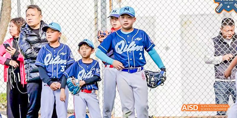Two young boys from International School of Dongguan stand on a baseball field in uniform, ready to practice catching and fielding drills.
