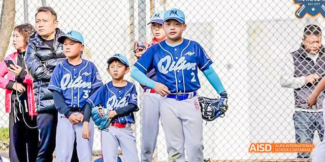 Two young boys from International School of Dongguan stand on a baseball field in uniform, ready to practice catching and fielding drills.