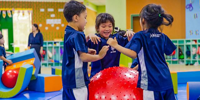 Three elementary students from International School of Dongguan share a joyful hug while playing with a large red ball during an indoor activity.