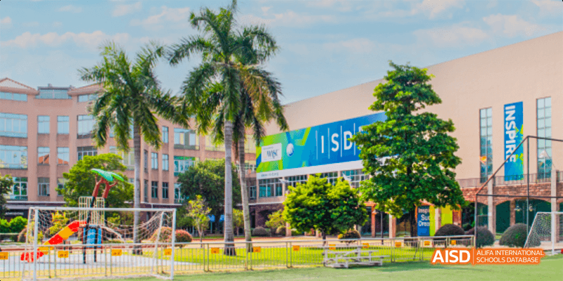 A view of the bright, modern campus building and palm trees at the International School of Dongguan, next to a green outdoor field.