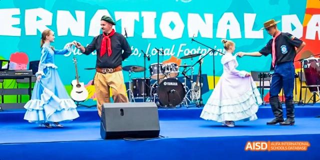 A male performer dressed in traditional or folk clothing with a red scarf on stage at an International School of Dongguan cultural or national event.