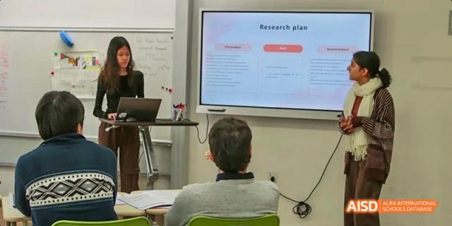 A teacher from JPED Academy, presenting a "Research Plan" on a large screen in a classroom.