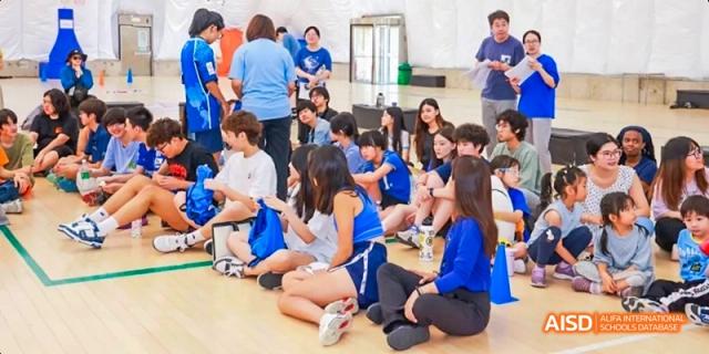 A large group of students from JPED Academy, sitting on the floor in a gym, listening to a teacher.