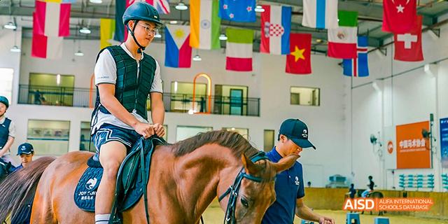 A student wearing a helmet while riding a brown horse with an instructor's guidance at Majestic International College.