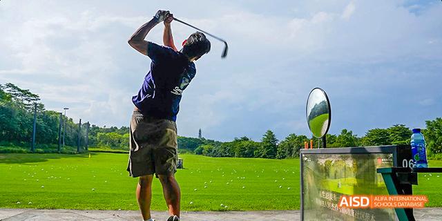 A student swinging a golf club, with a wide green field and a golf ball visible, at Majestic International College.