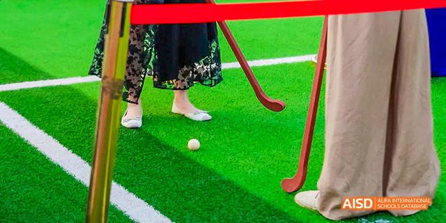 A close-up shot of a student hitting a white ball with a red field hockey stick on an artificial turf field at Majestic International College.