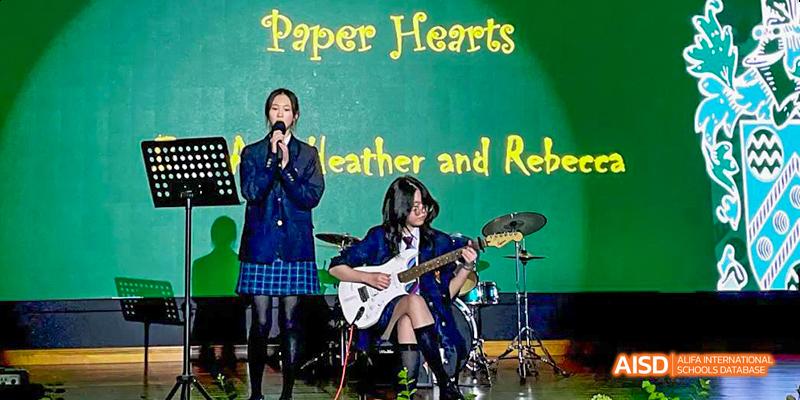 Female students are performing on a stage, with one singing and another playing an electric guitar at Malvern College Chengdu.