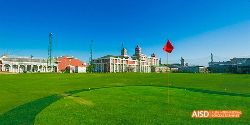 The main academic building of Beijing Aidi School, a red and white structure with a large grassy field in front of it under a clear blue sky