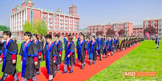 Beijing Aidi School students in graduation gowns walking on a red carpet in front of the school's main building.