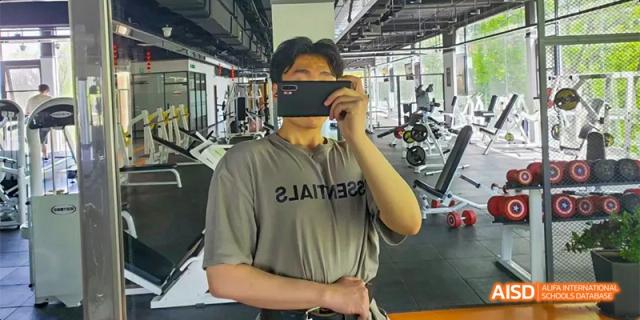 A male student at Beijing Aidi School wearing a gray shirt, taking a photo of himself in a gym.