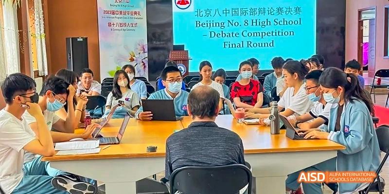 A debate competition final round at Beijing No.8 High School, showing students and a judge seated around a table.