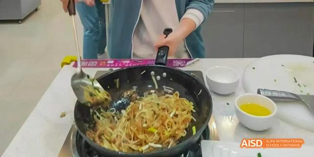Close-up of a stir-fry being prepared in a black pan, showing noodles and vegetables, as part of a Beijing No.8 High School cooking class.