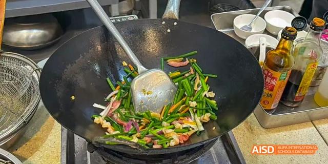 Vegetables and spices being stir-fried in a large wok with a metal spatula during a Beijing No.8 High School cooking activity.