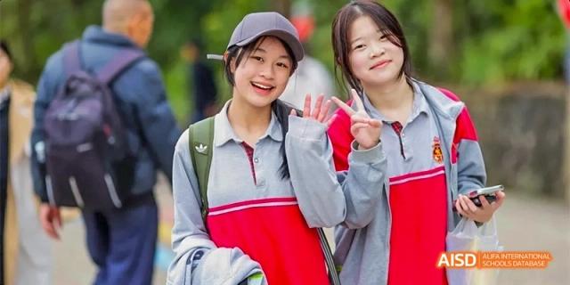 Two female students, one wearing a gray and red uniform and a cap, smiling and waving outdoors at DongGuan Laguna Verona Foreign Language School.