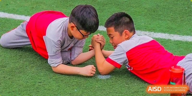Two male students arm wrestling on a grassy field, wearing school uniforms with red accents, at DongGuan Laguna Verona Foreign Language School.