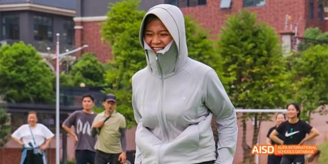 A female student wearing a light gray zipped hoodie with the hood up, smiling outdoors on campus at DongGuan Laguna Verona Foreign Language School.