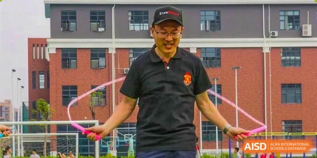 An adult male teacher in a black polo shirt and cap smiling while exercising with a jump rope outdoors on the grounds of DongGuan Laguna Verona Foreign Language School.