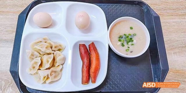 A school cafeteria tray with a breakfast of dumplings, bacon strips, an egg, and a small bowl of porridge at DongGuan Laguna Verona Foreign Language School.