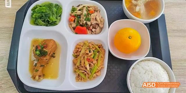 A school lunch tray with a mix of stir-fried vegetables and meat, a piece of fruit, and a cup of liquid, served in the cafeteria at DongGuan Laguna Verona Foreign Language School.