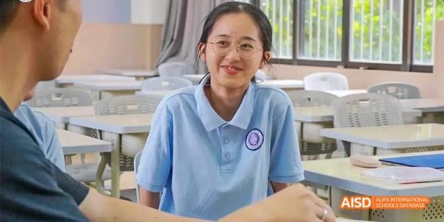 A close-up of a smiling female student wearing a light blue school uniform and glasses, seated at her desk in a classroom at GDUFS International School Guangzhou.