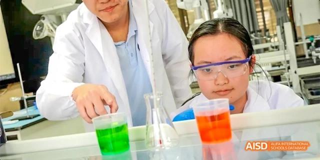 Students in lab coats and safety glasses conducting a chemistry experiment with brightly colored liquids in beakers at GDUFS International School Guangzhou.