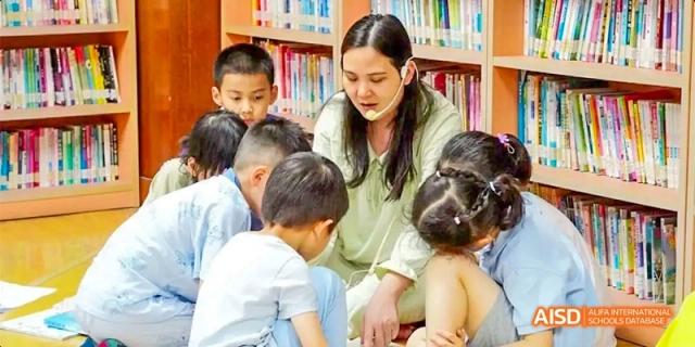 A teacher reading a book to a group of young students seated on the floor in the children's section of the library at GDUFS International School Guangzhou.