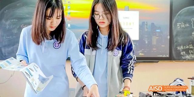 Two female students wearing school uniforms and glasses working together on a computer or digital task at a table at GDUFS International School Guangzhou.
