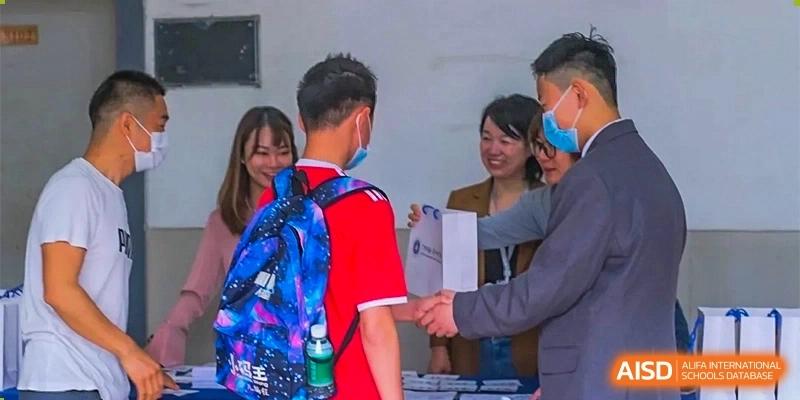 A male student with a blue backpack shaking hands with a teacher or staff member during a greeting or registration at GDUFS International School Guangzhou.