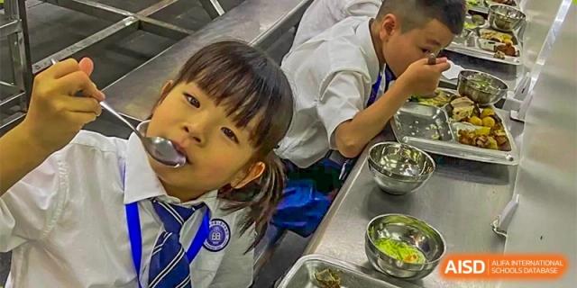 A close-up of a young female student with a spoon, looking at the camera while eating a meal in a dining area at GDUFS International School Guangzhou.