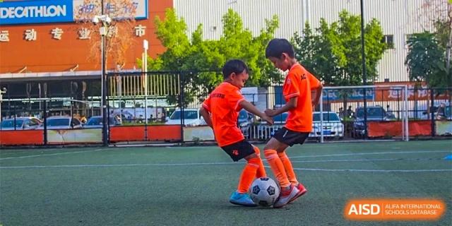 Two young male students in orange uniforms playing soccer on a green turf field, with one student controlling the ball at GDUFS International School Guangzhou.