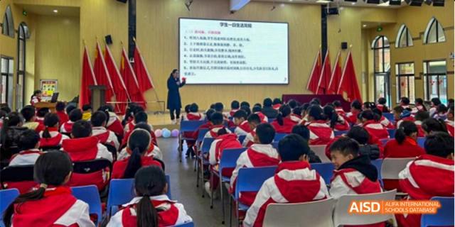 A Guiyang Prime School classroom or lecture hall with students in red and blue uniforms facing a screen where a teacher is giving a lesson.