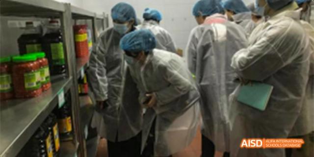 Guiyang Prime School students or staff in protective gear (smocks and hairnets), observing or working in a factory or clean room environment.