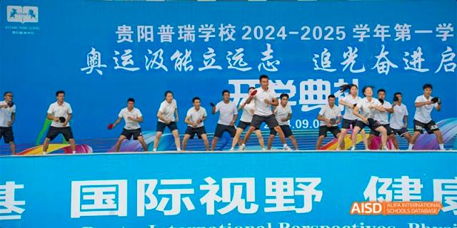 Guiyang Prime School students or staff performing a routine on a stage visible in front of a large blue banner that displays the school name and event details.