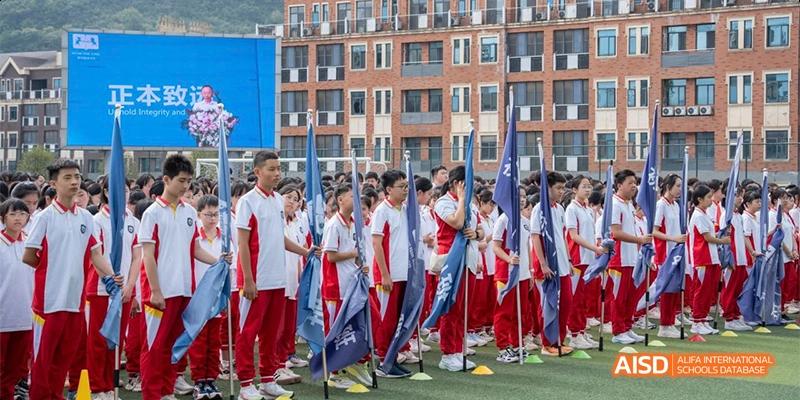 Guiyang Prime School students standing in formation outdoors, holding flags or banners during a school event or sports day, with school buildings in the background.